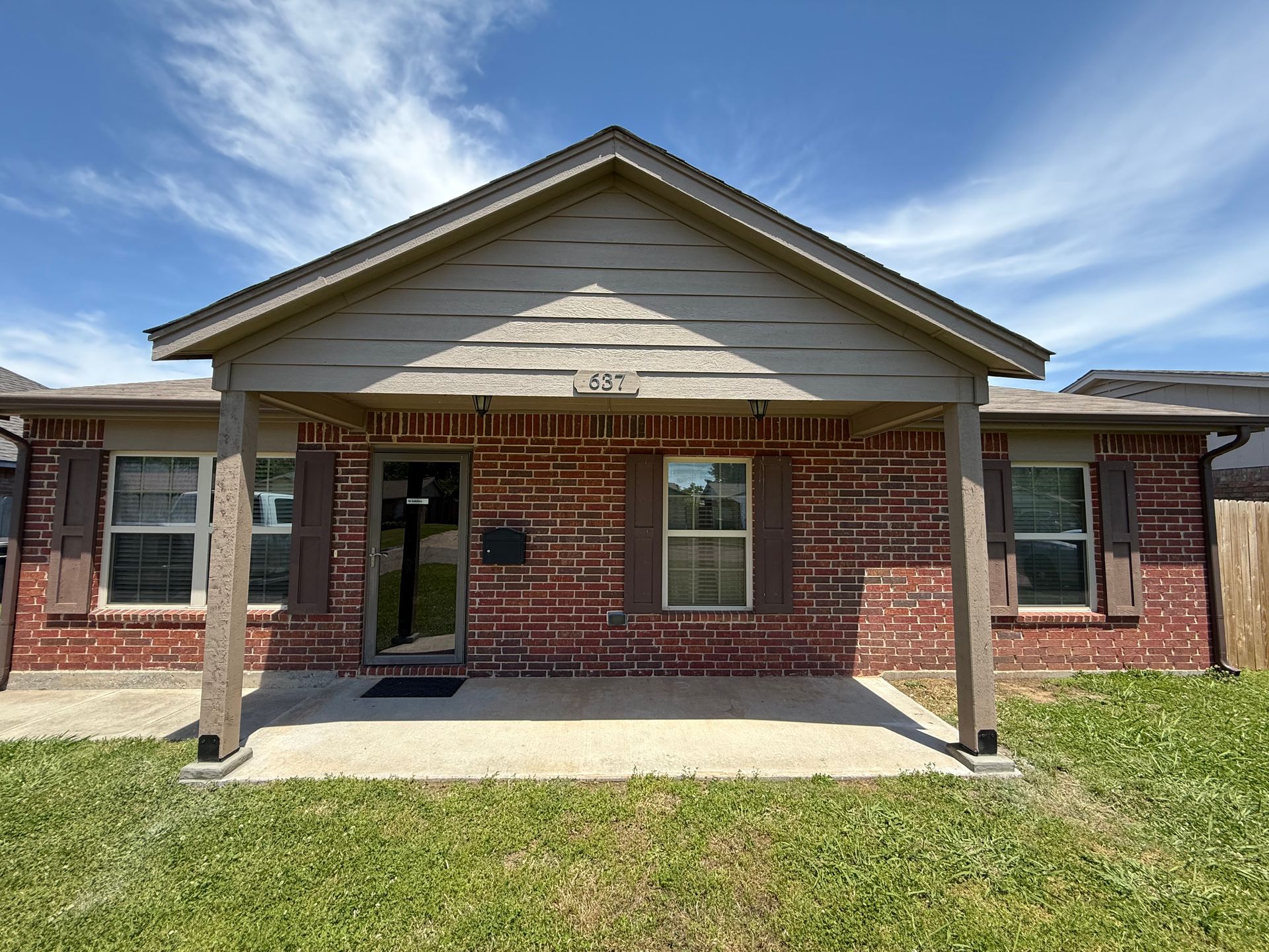 Red brick house with porch, brown shutters, and green lawn under a blue sky.