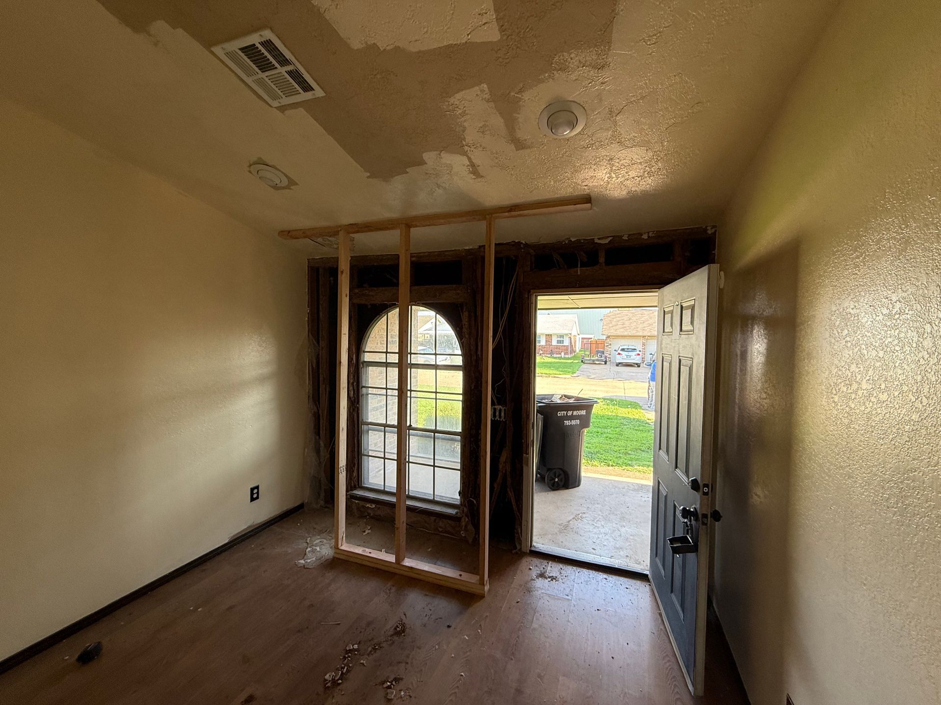 Interior view of a damaged entryway. Wood framing surrounds an arched window and open doorway to the outside.