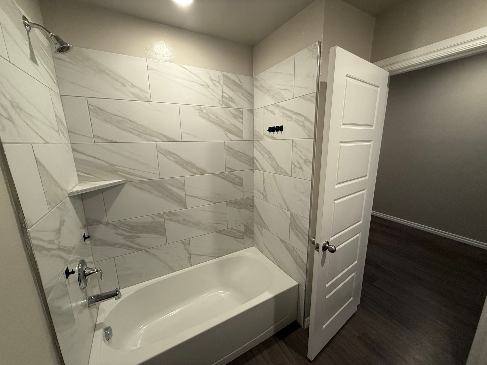Bathroom with white tub, marble tile shower, and white door leading to a dark floor room.