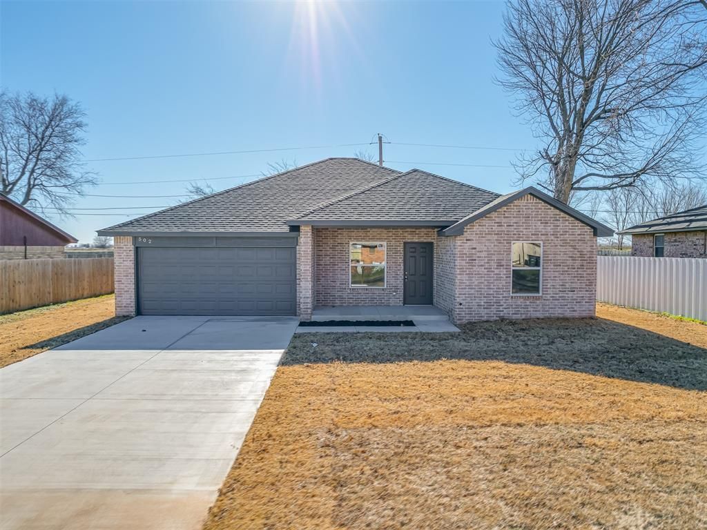 Single-story brick house with a gray garage door, concrete driveway, and brown roof under a sunny sky.