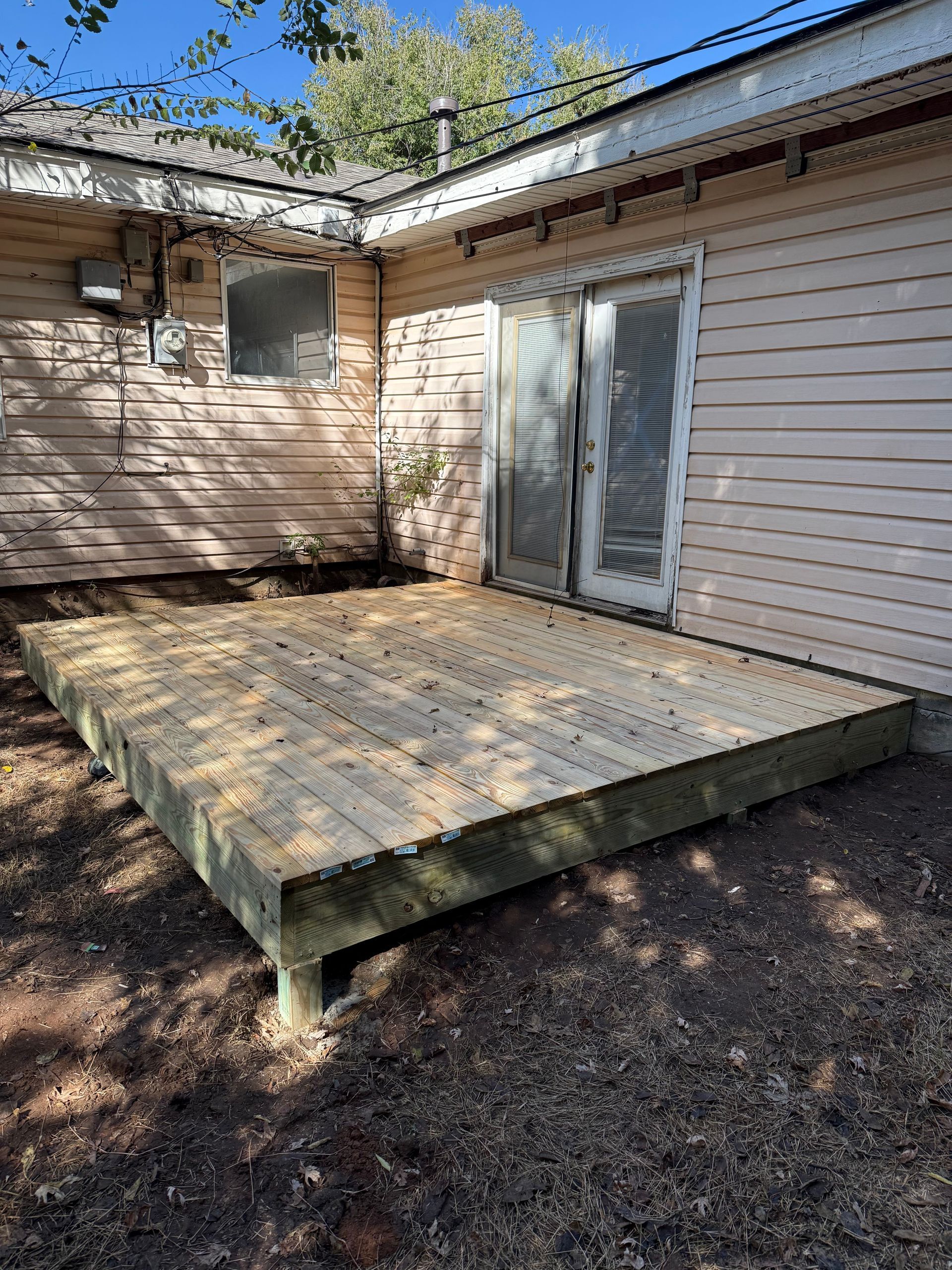 Wooden deck outside a building with light pink siding and glass doors. Ground is bare dirt.