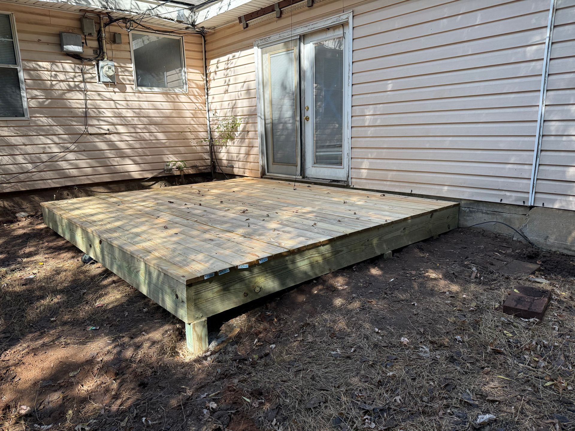 Wooden deck outside a house with a sliding glass door. The ground is dirt.