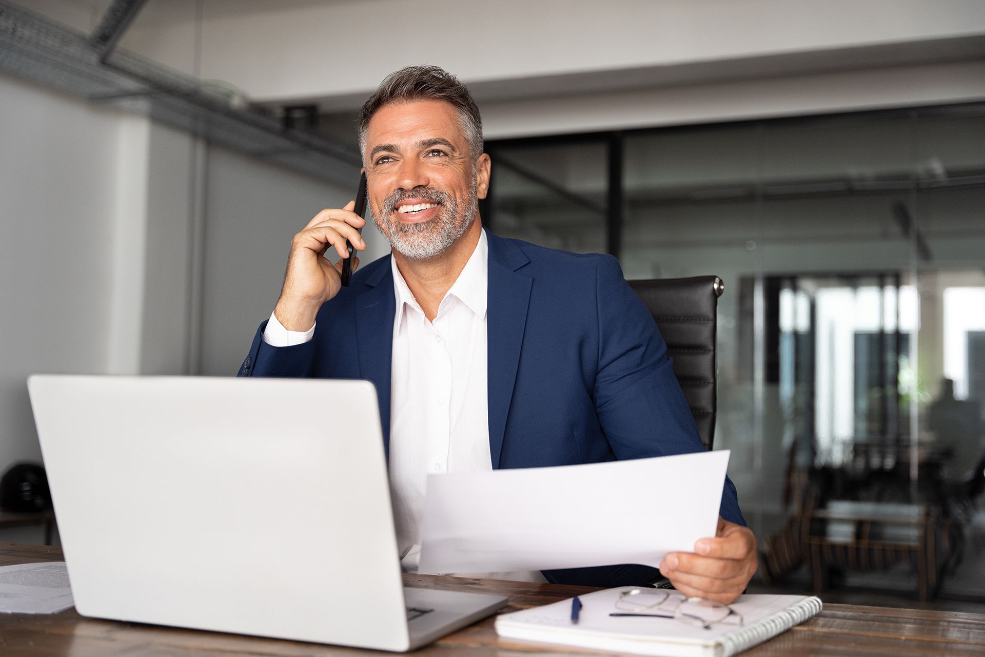 A man in a suit is sitting at a desk talking on a cell phone.