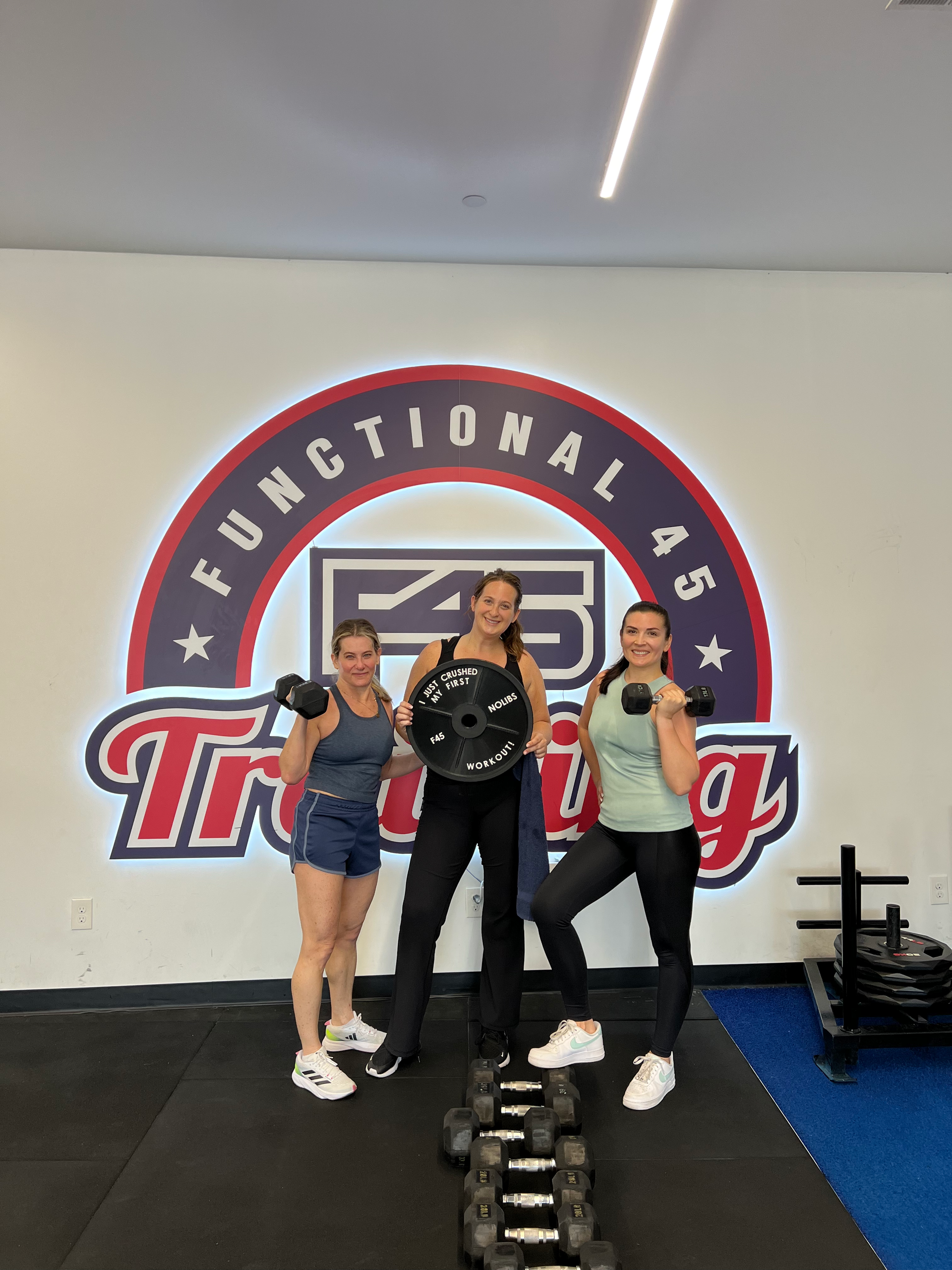 Three women are posing for a picture in front of a functional as training logo