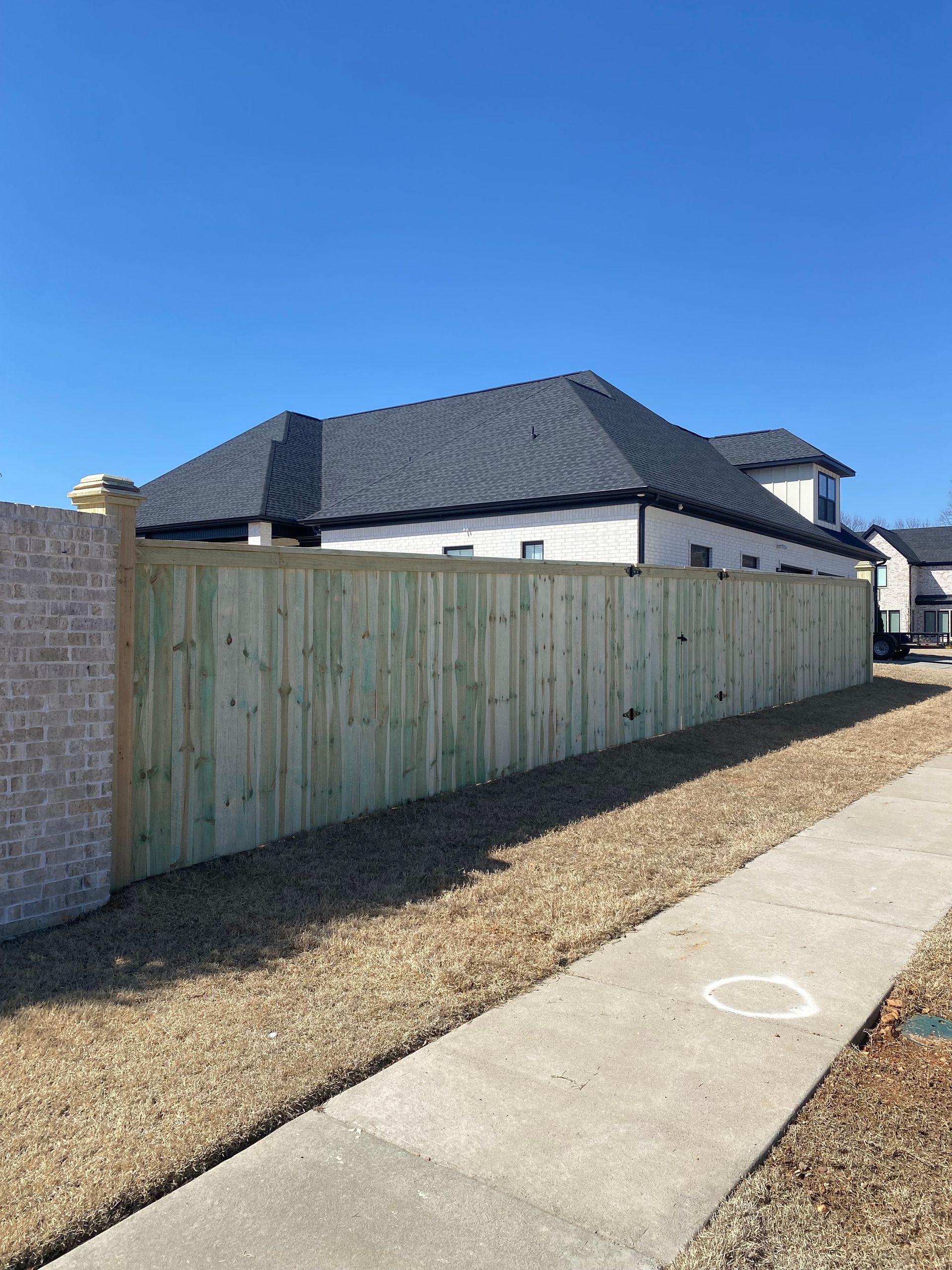 A wooden fence next to a sidewalk and gravel, with a house and clear blue sky in the background.
