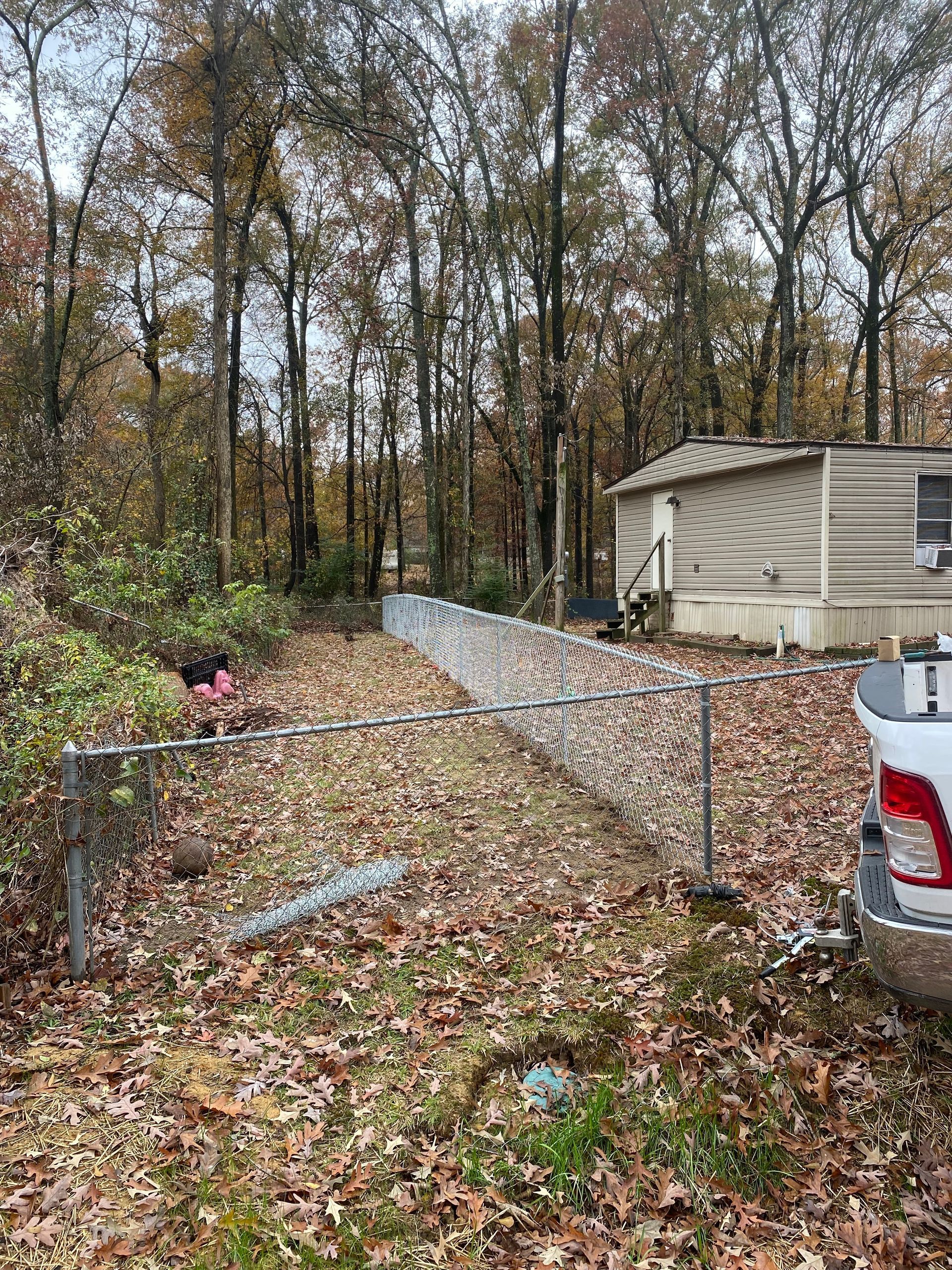 Chain-link fence in wooded area near a small building and white truck with fallen leaves on the ground.