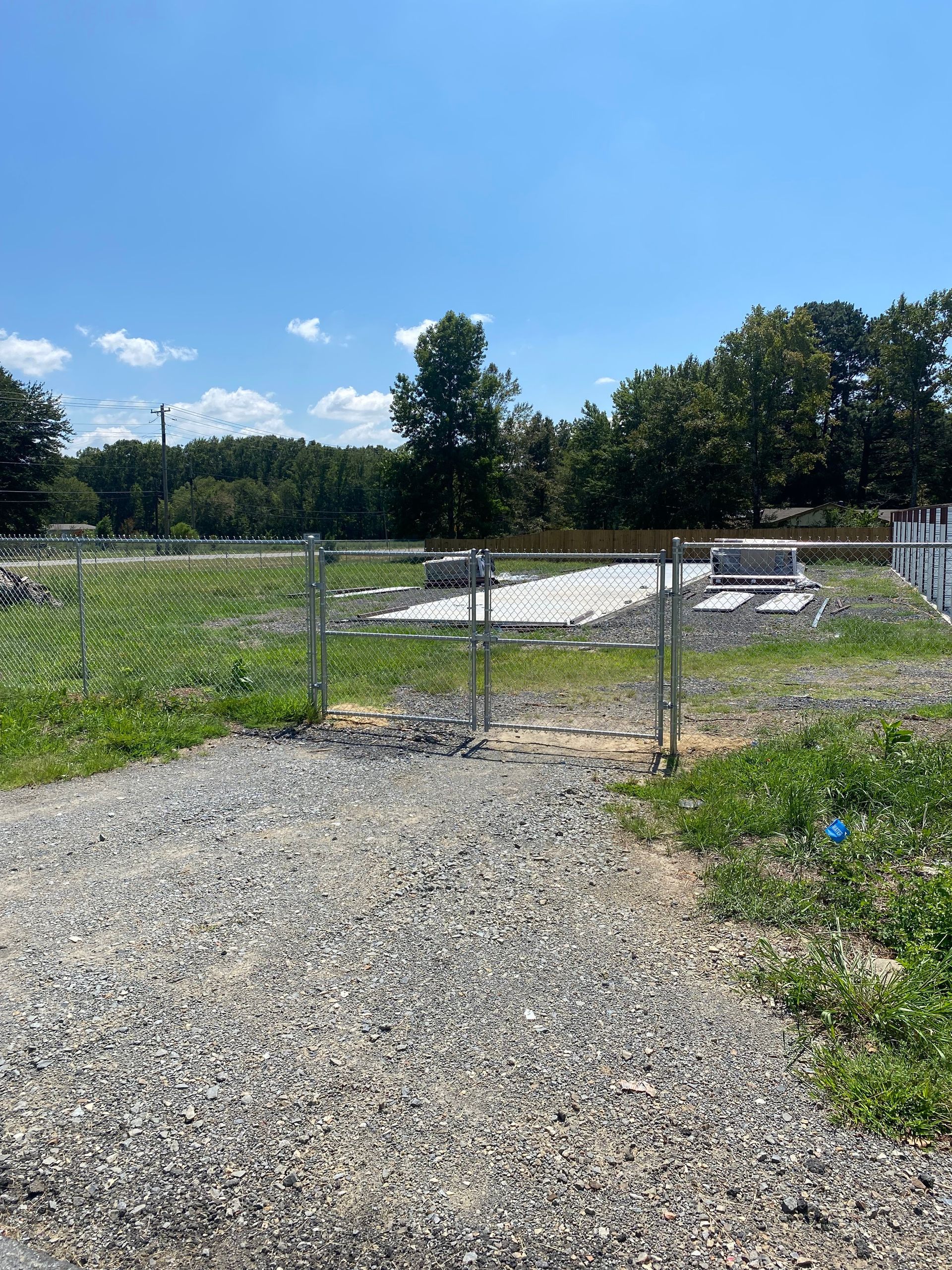 Gravel path leading to a chain-link fence with a gate open to a grassy area under a blue sky.