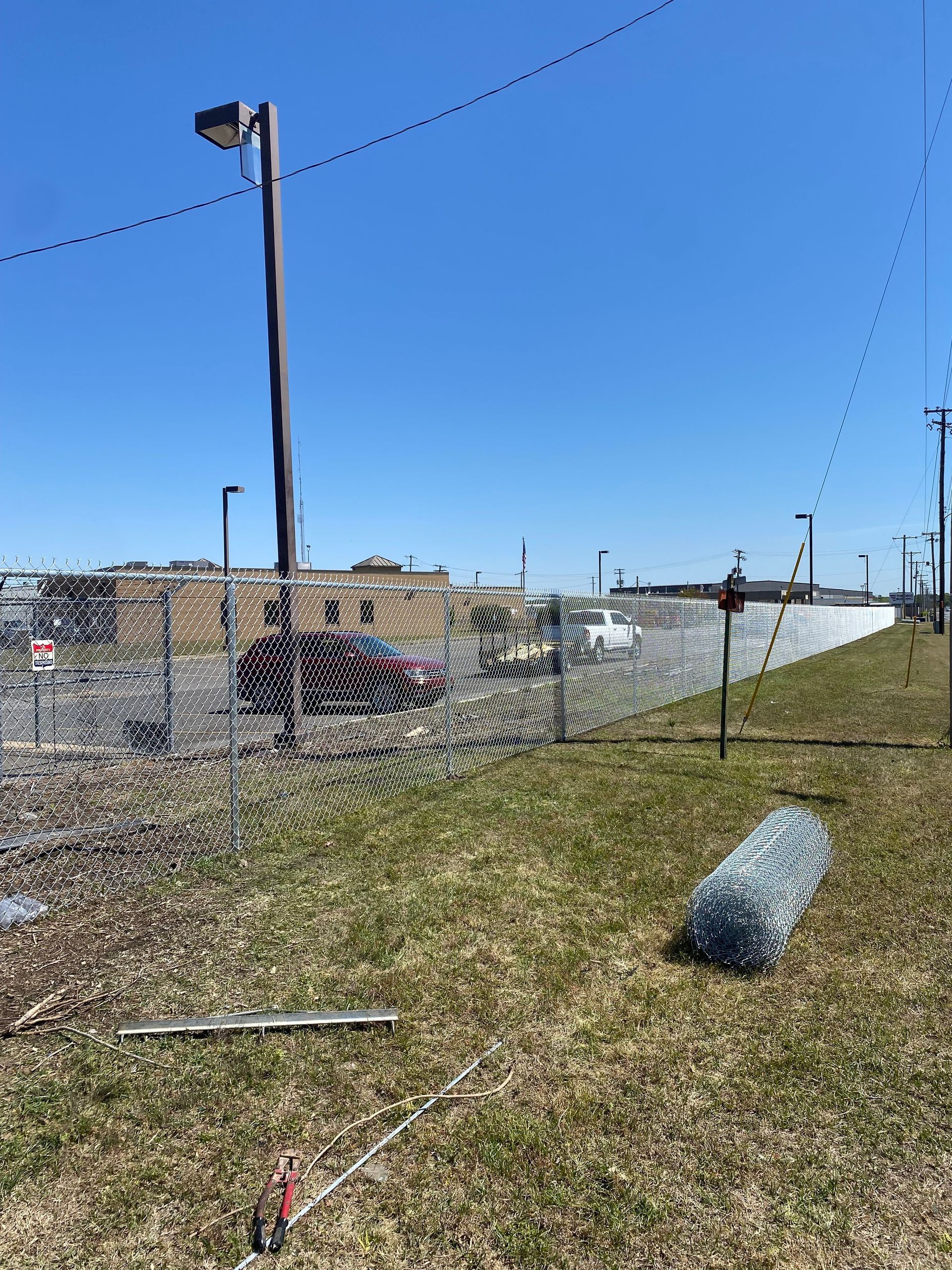 Chain-link fence in front of a building on a sunny day; grass in foreground with a rolled-up fence segment.