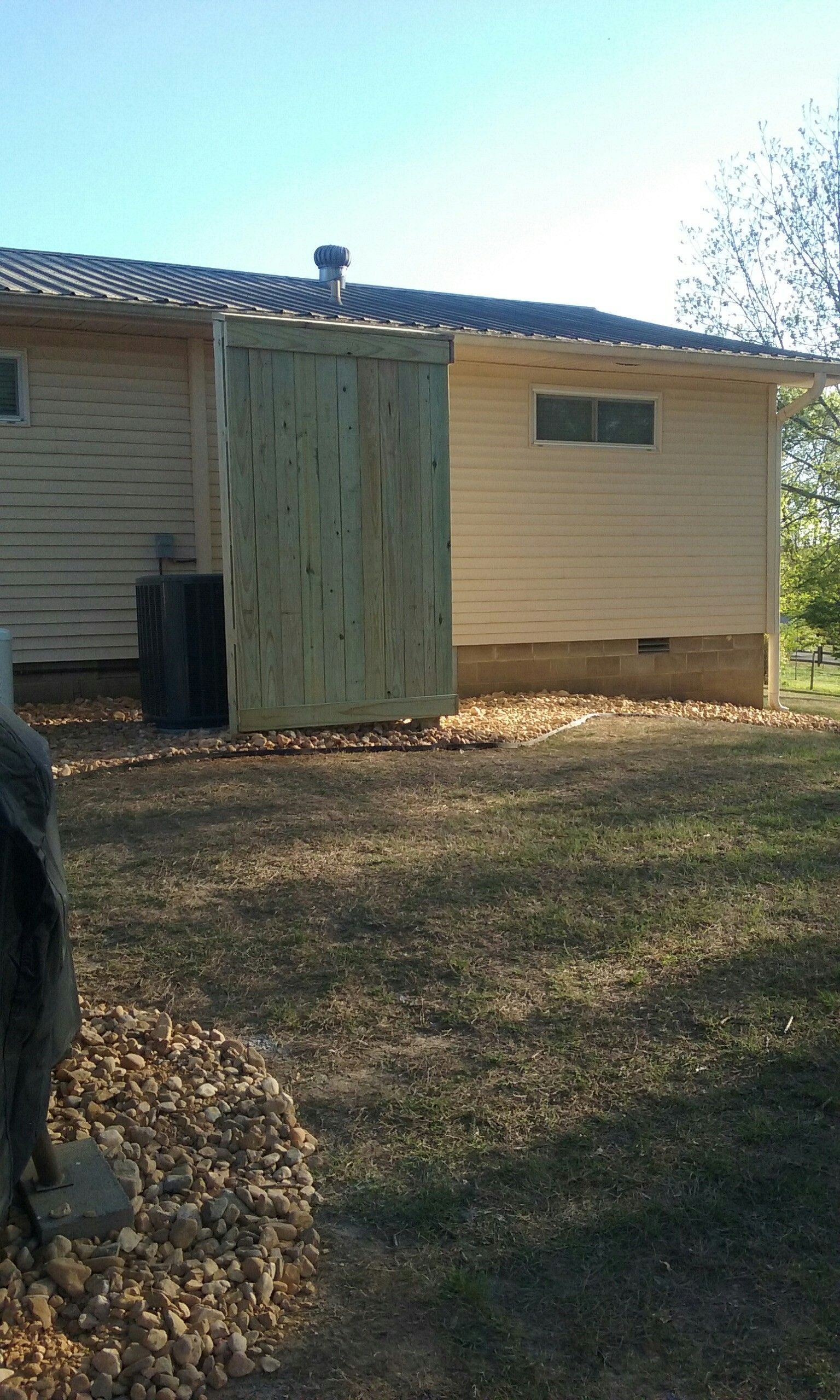 A wooden structure next to a beige house, obscuring an air conditioning unit. Autumn yard.