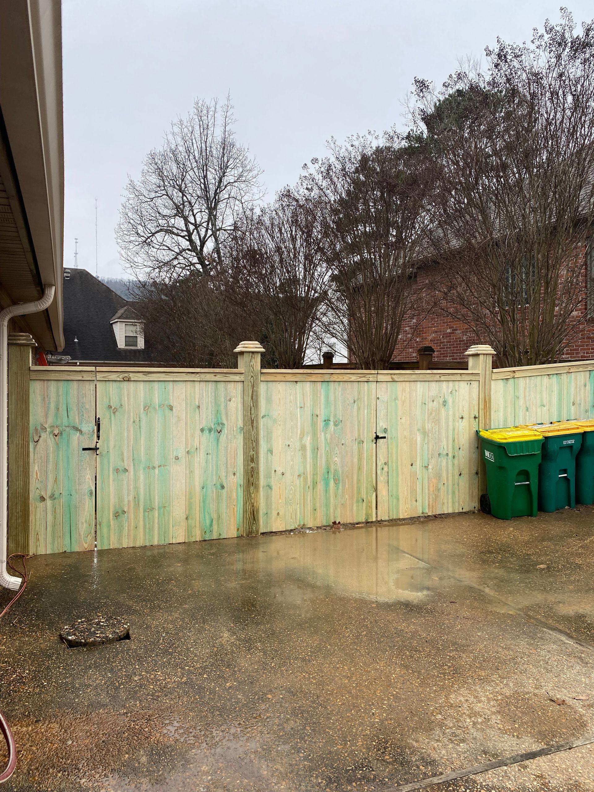 Wooden fence with gate, green trash cans. Rainy outdoor setting.