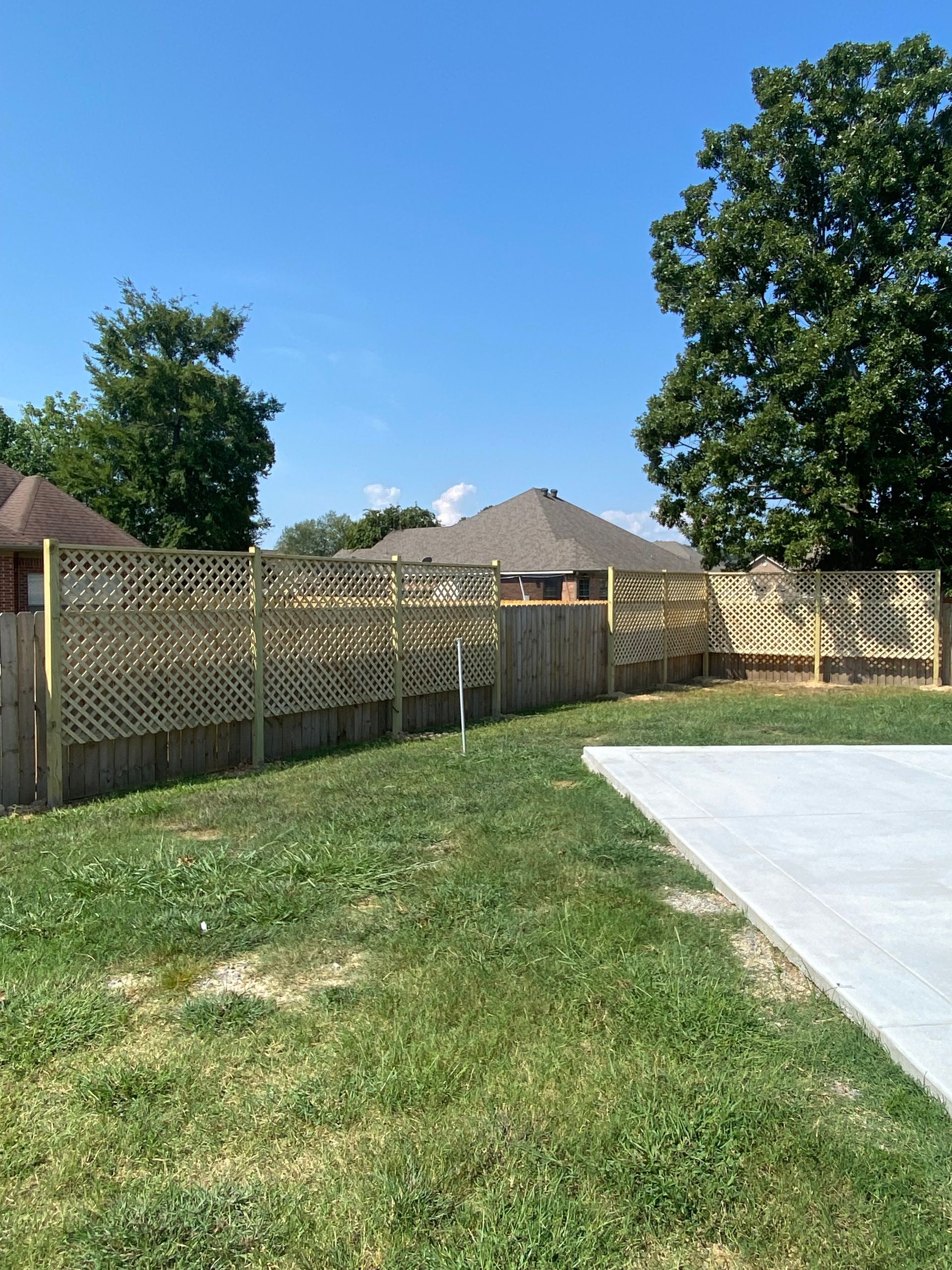 Wooden fence with lattice top in a backyard, sunny day. Concrete slab in the foreground.