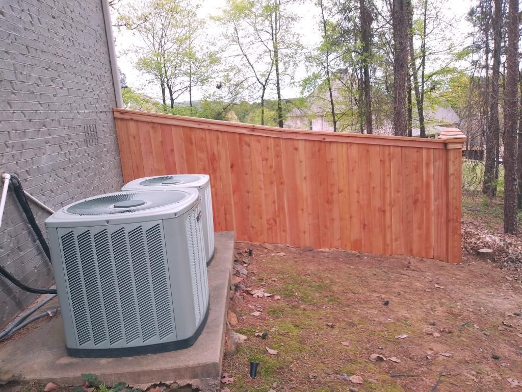 Two air conditioning units next to a wooden privacy fence in a wooded area.