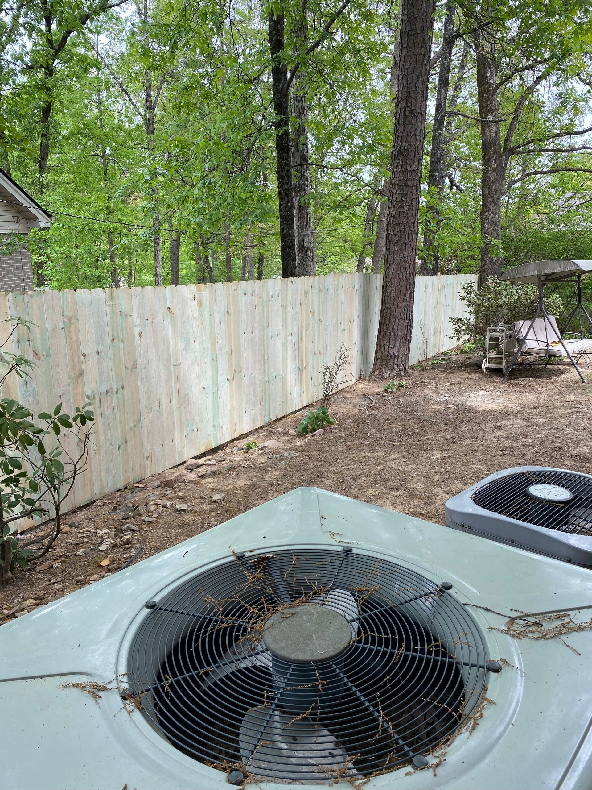 A new wooden fence next to trees in a backyard, with an air conditioning unit in the foreground.