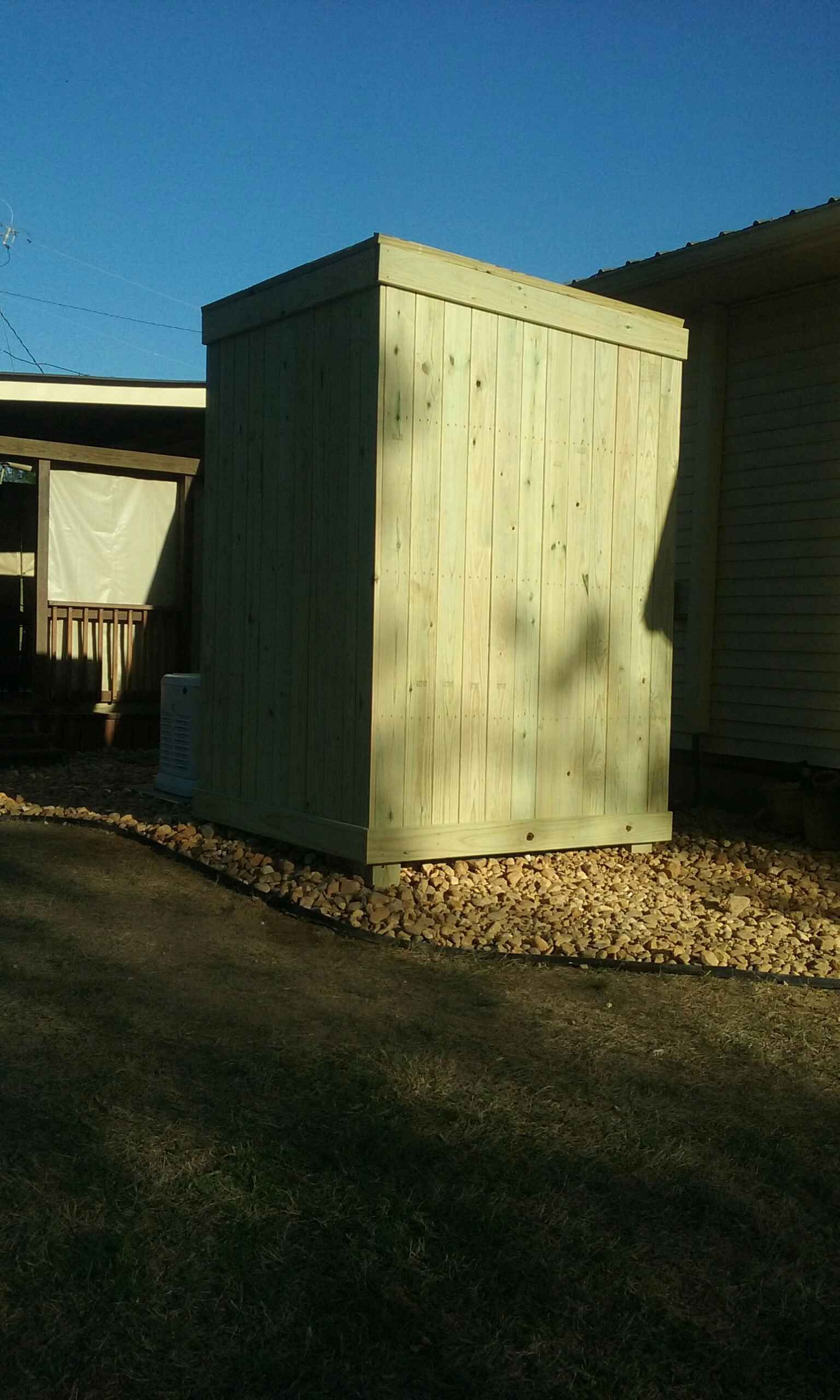 A light green, wooden utility box sits on gravel next to a building on a sunny day.
