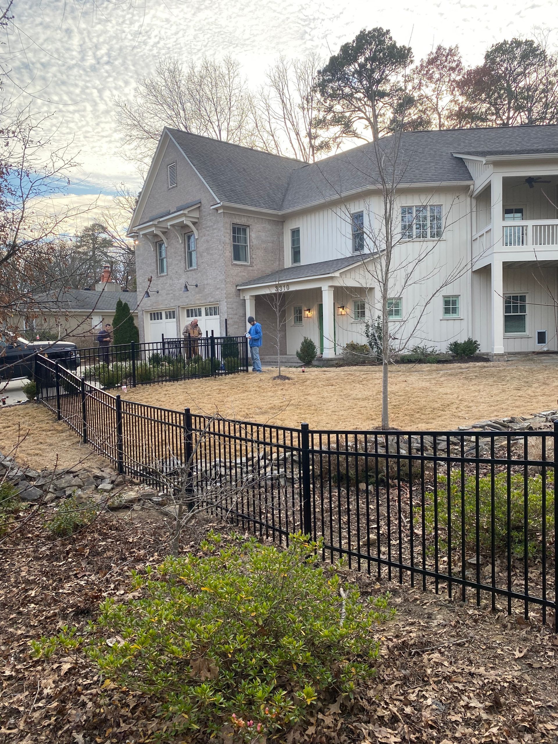 Two-story house with light brick and siding, black fence in front. Bare yard, two people near house.