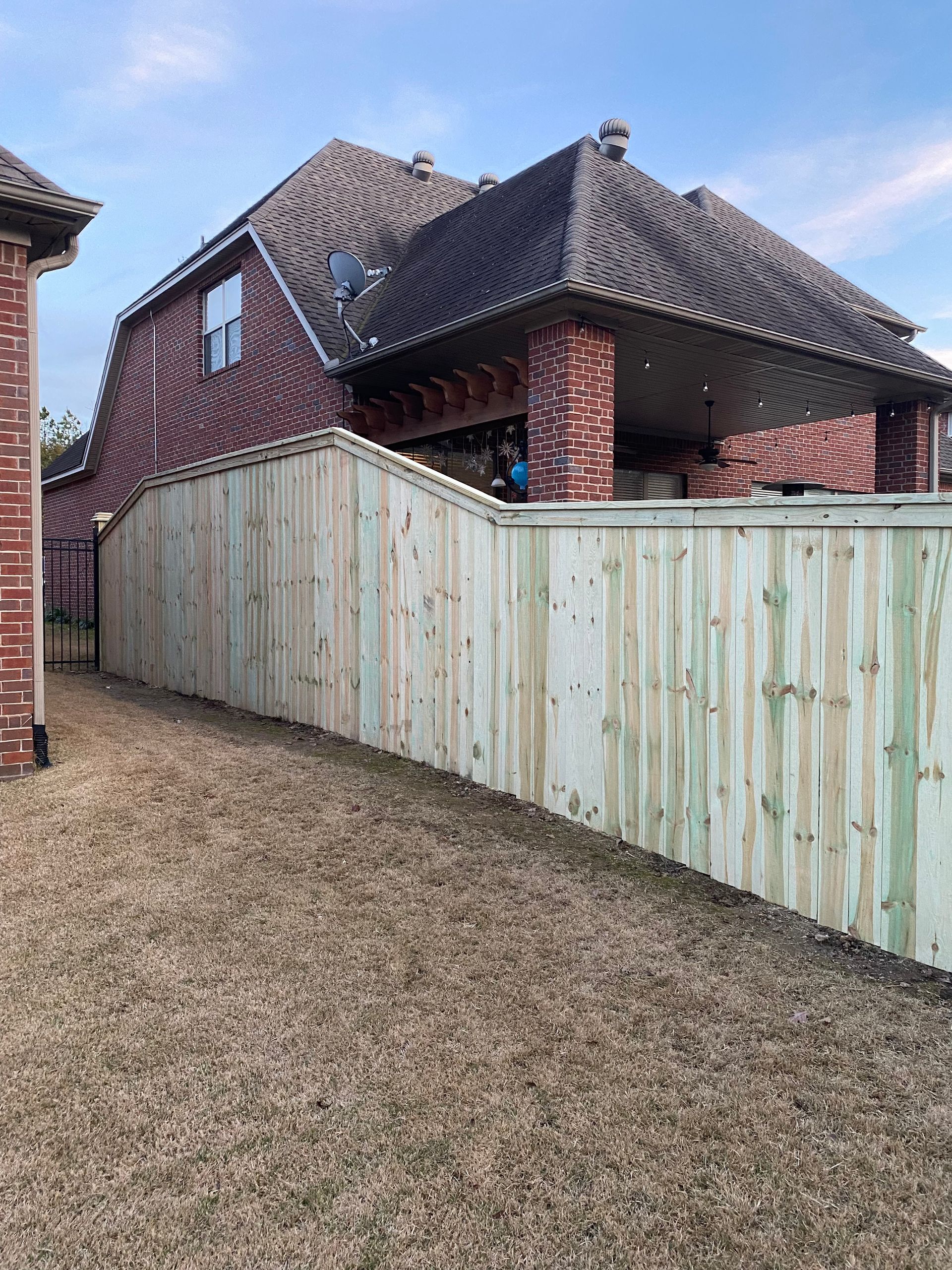 Wooden fence along the side of a brick house, partially covering a porch; brown yard.