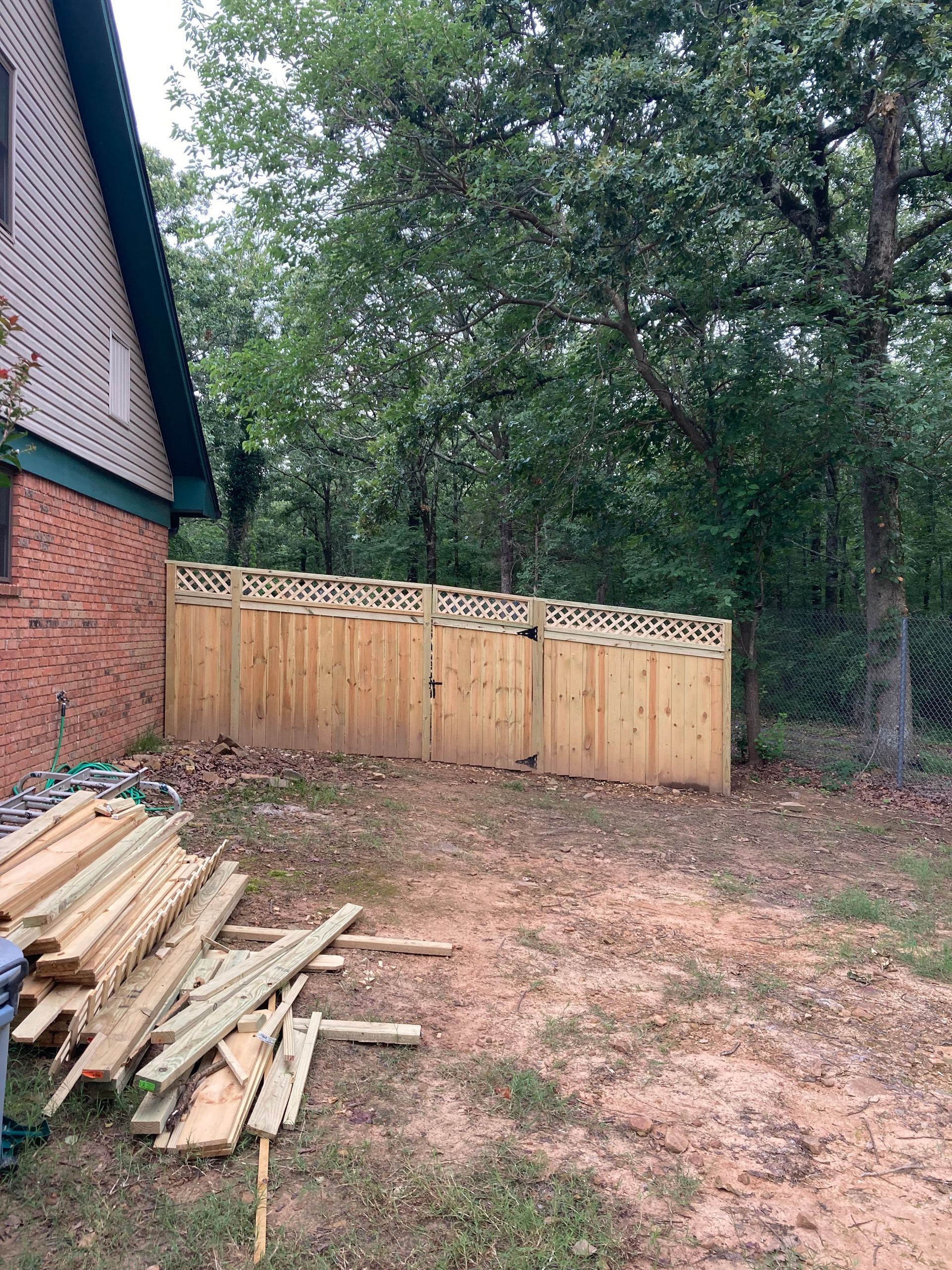 A wooden fence with decorative top, next to a brick building and trees, with lumber in the foreground.