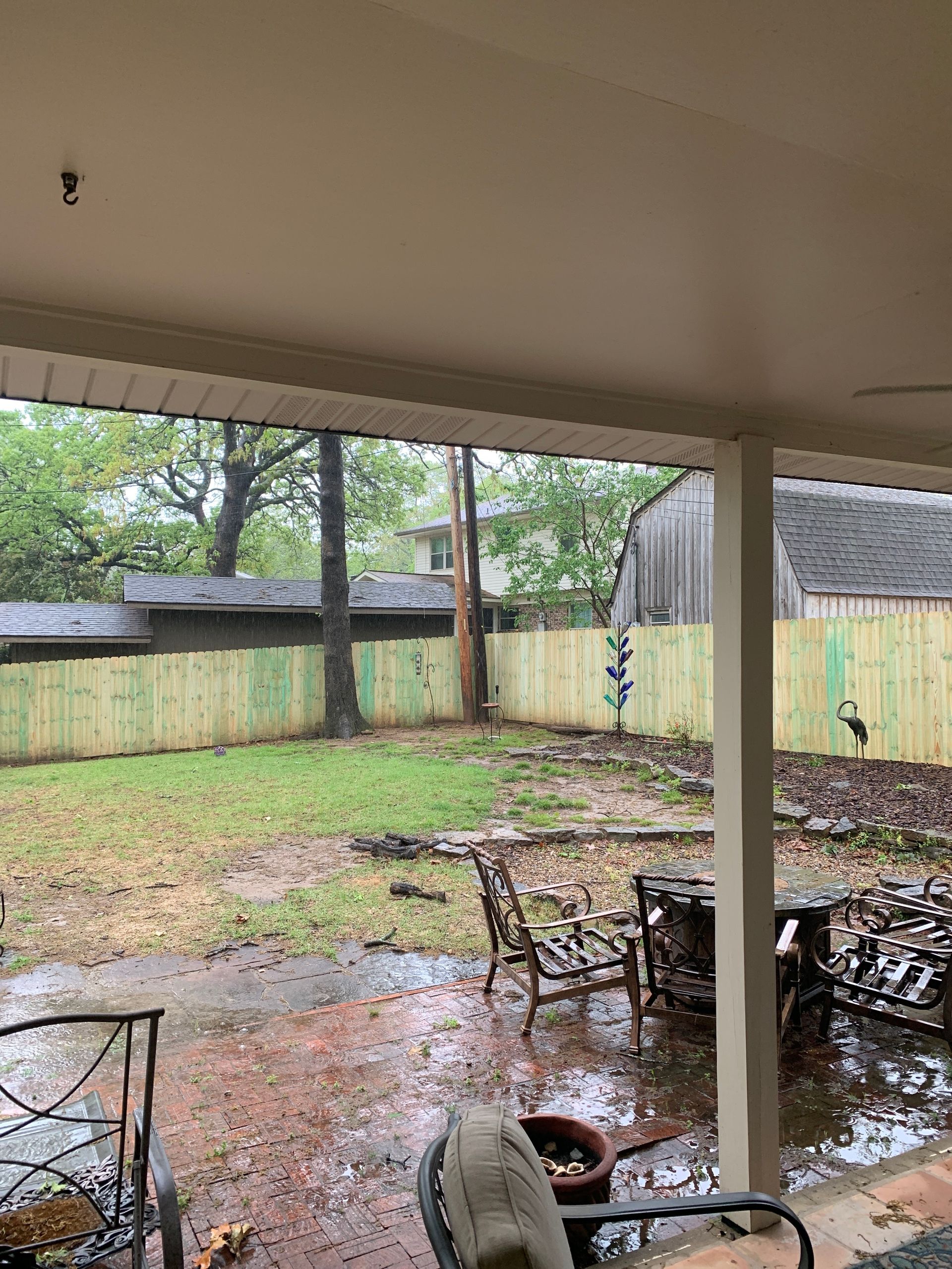 Rainy backyard view from a porch. A wooden fence surrounds a soggy yard, with a tree and shed in the distance.
