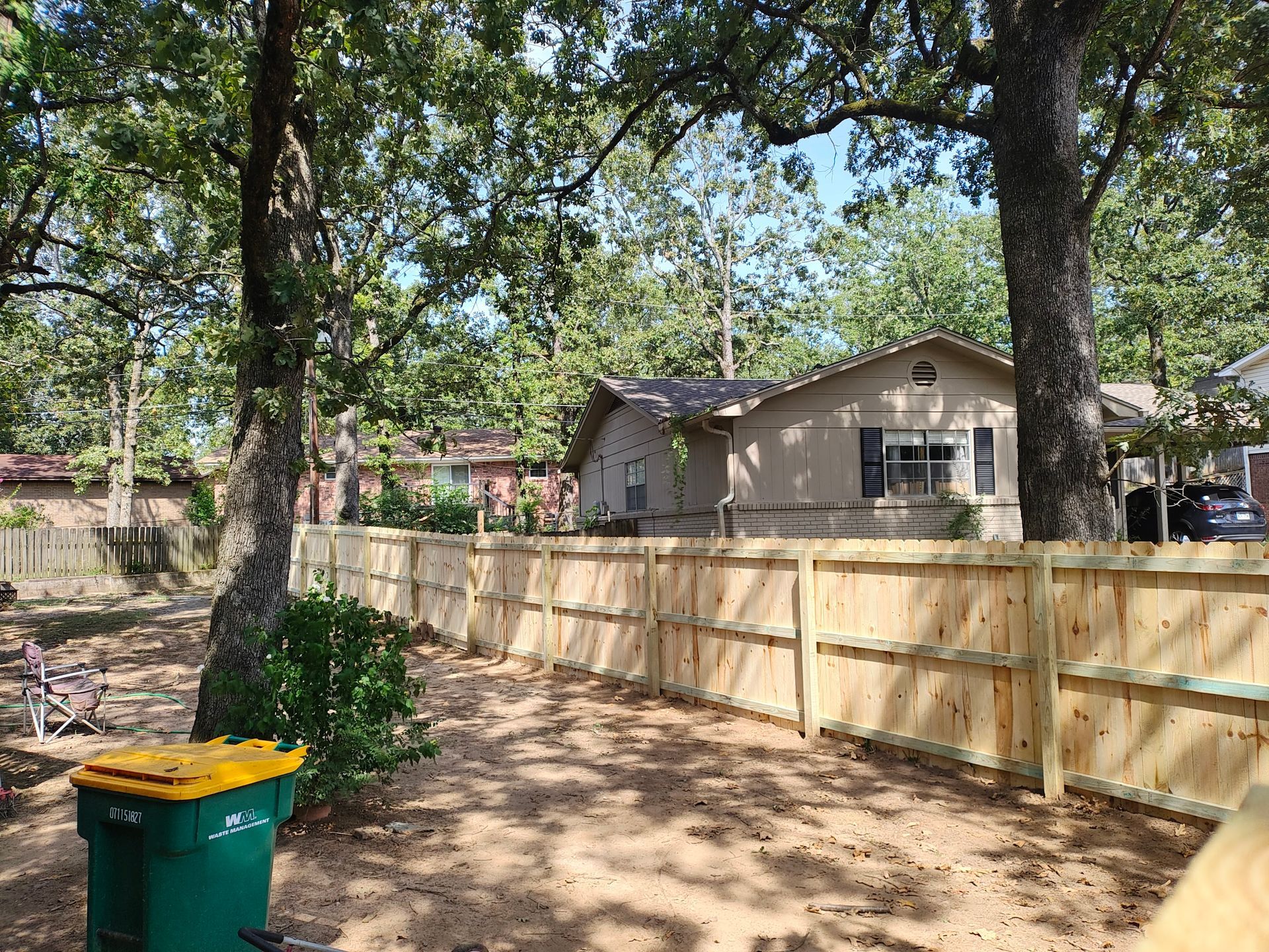 Wooden fence in a backyard with trees and a house in the background. A green trash bin is in the foreground.