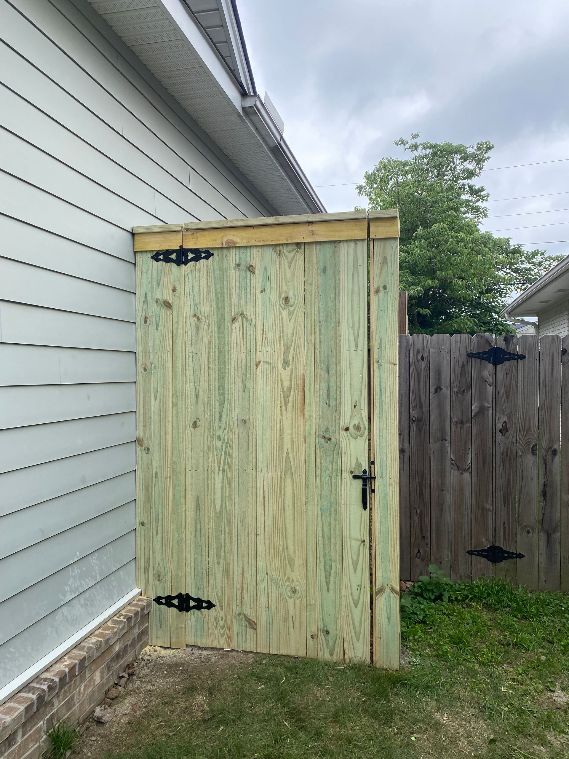 Wooden storage enclosure attached to a house with a door, black hinges, and handle; beside a fence.