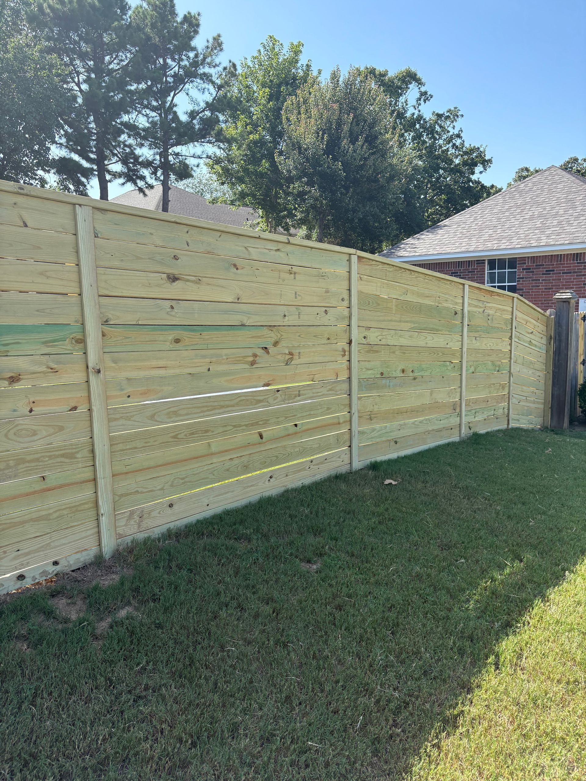 Wooden horizontal slat fence in a grassy yard, partially obscuring a house and trees.