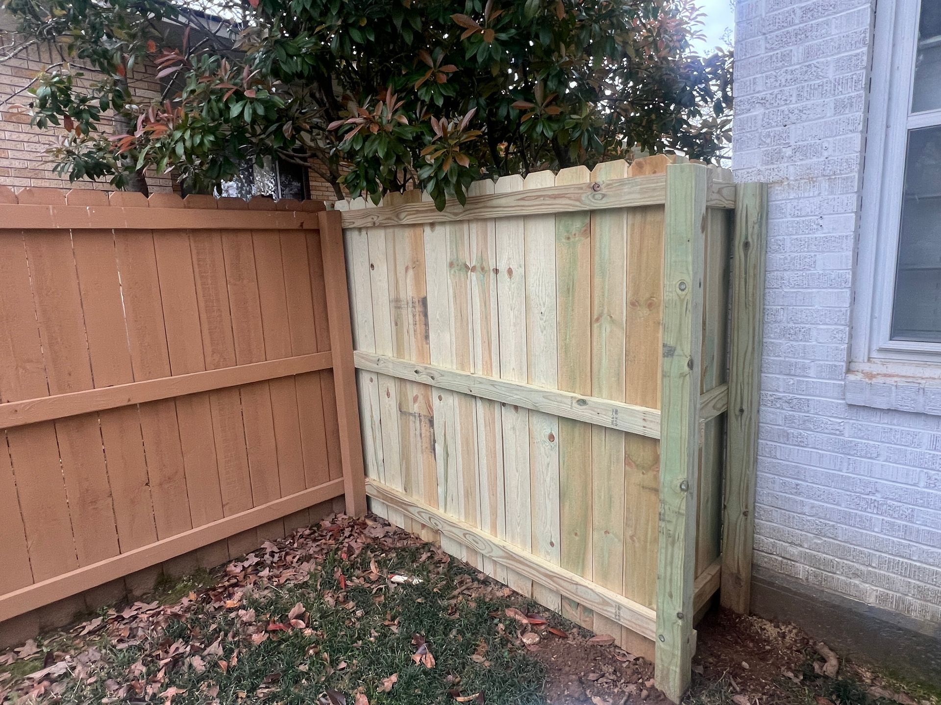 New wooden fence section against a brick wall, next to a brown fence and a tree.