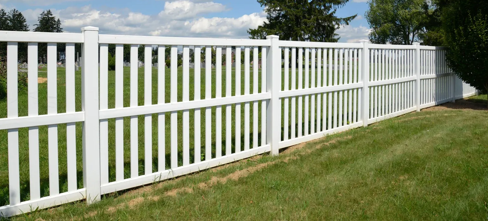White picket fence on green lawn under a blue sky with clouds.