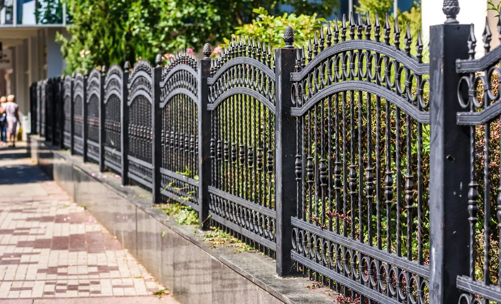 Black wrought iron fence along a brick sidewalk, with lush greenery behind it.