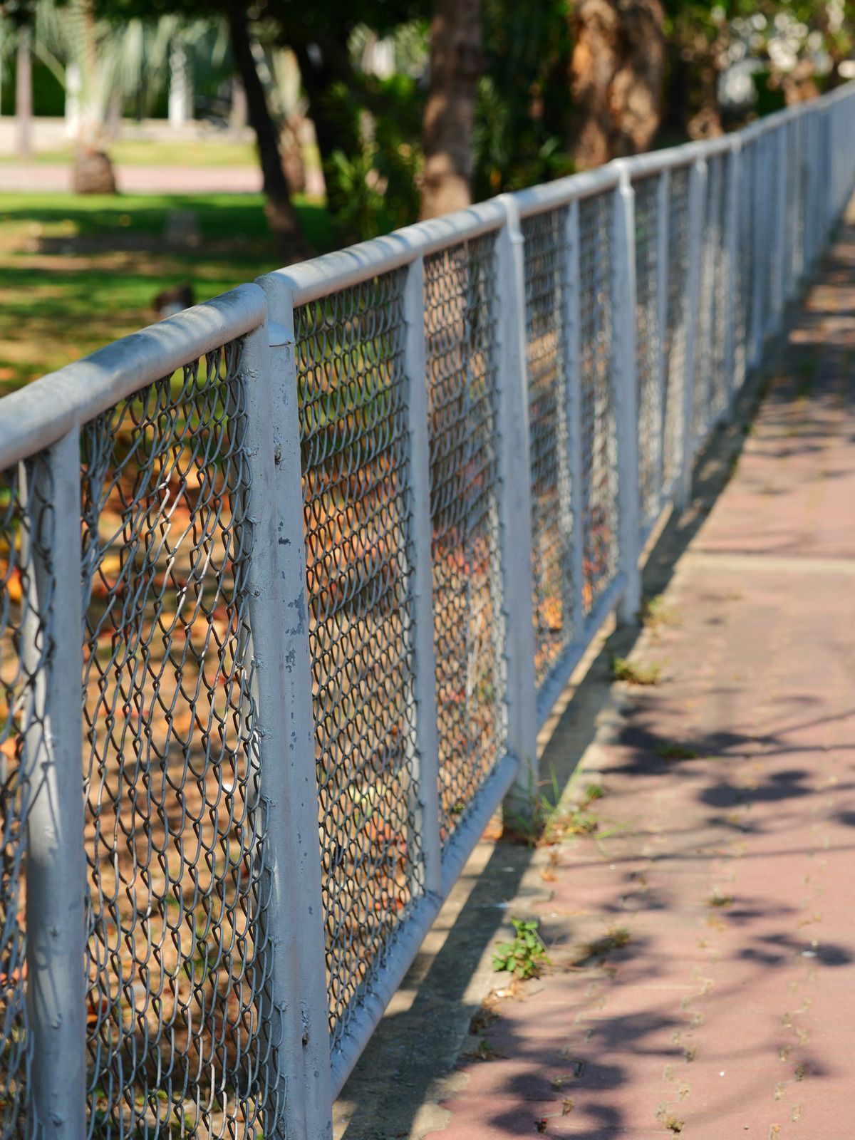 A white metal fence with wire mesh runs along a red brick path, with trees in the background.