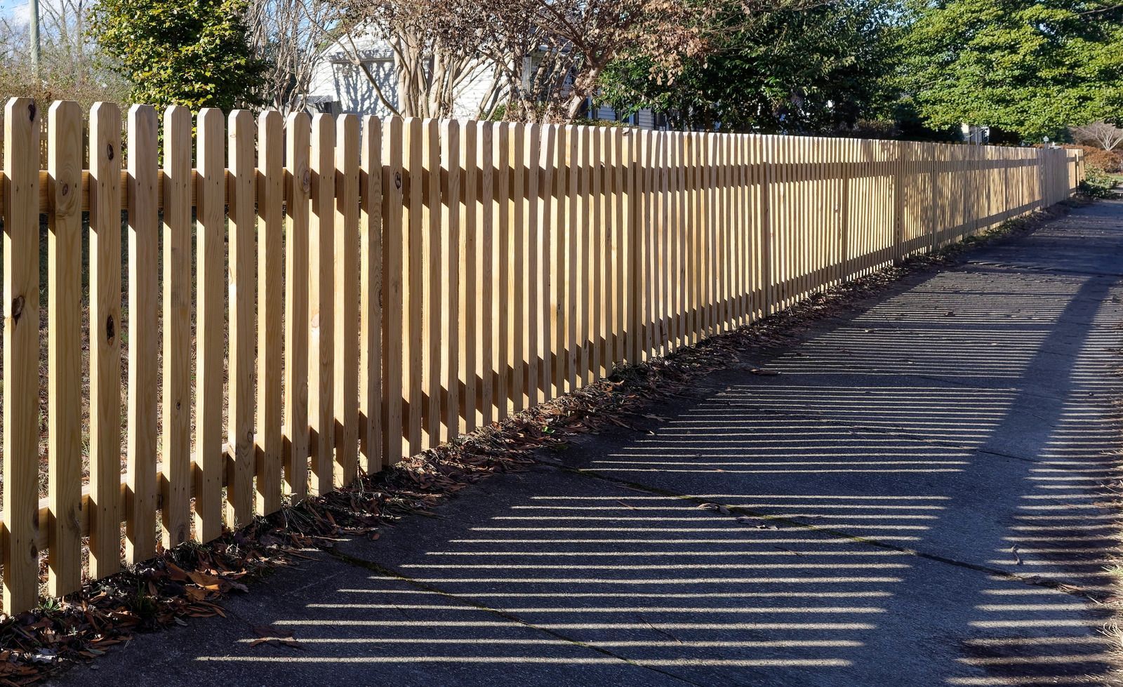 Wooden picket fence casting shadows on a paved surface, with sunlight and trees in the background.