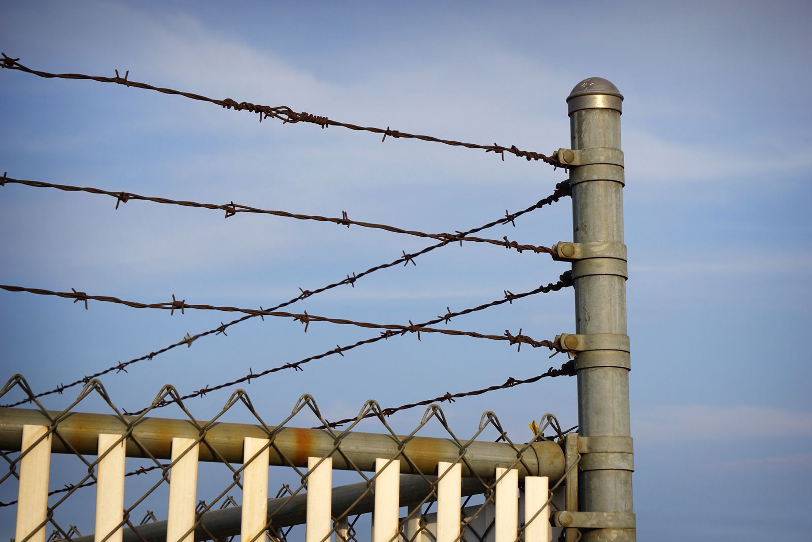 Barbed wire fence atop a chain-link fence, attached to a metal post, against a blue sky.