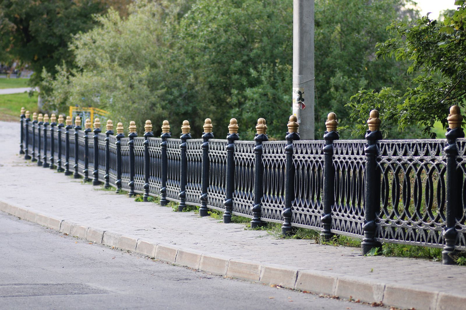 Black decorative metal fence with gold accents along a sidewalk next to a street, with trees in the background.