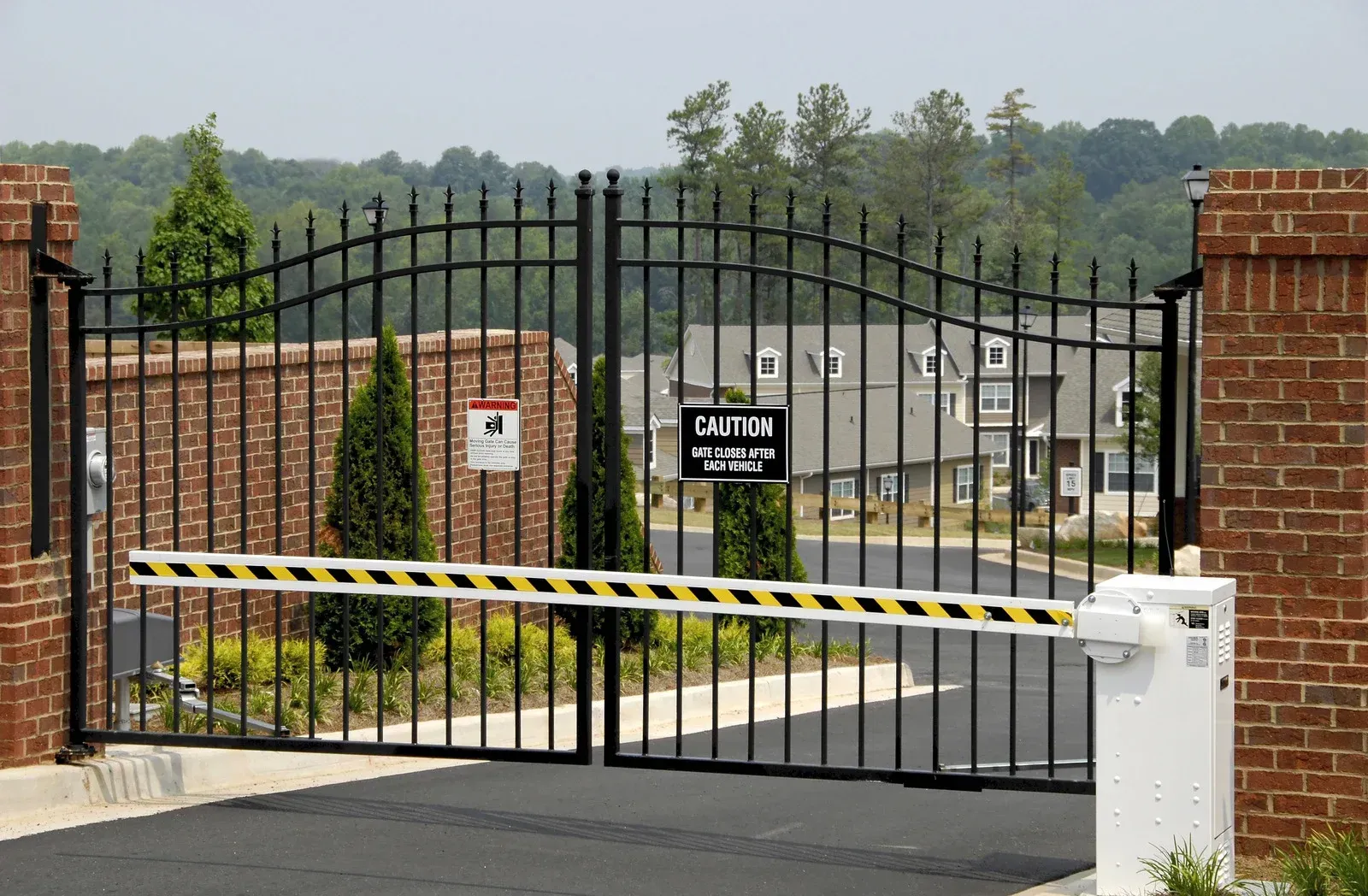 Black iron gate with a yellow and black striped arm barrier, brick pillars, and a sign.