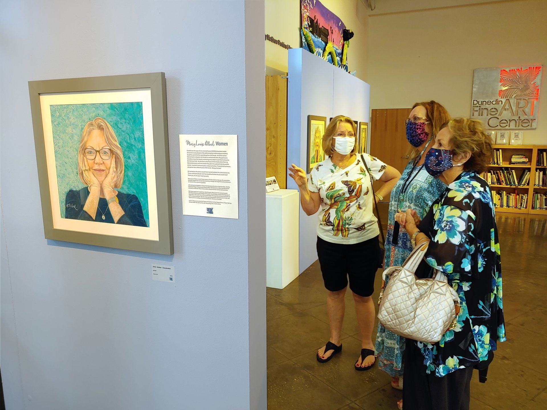 Three women wearing face masks are looking at a painting of a woman.