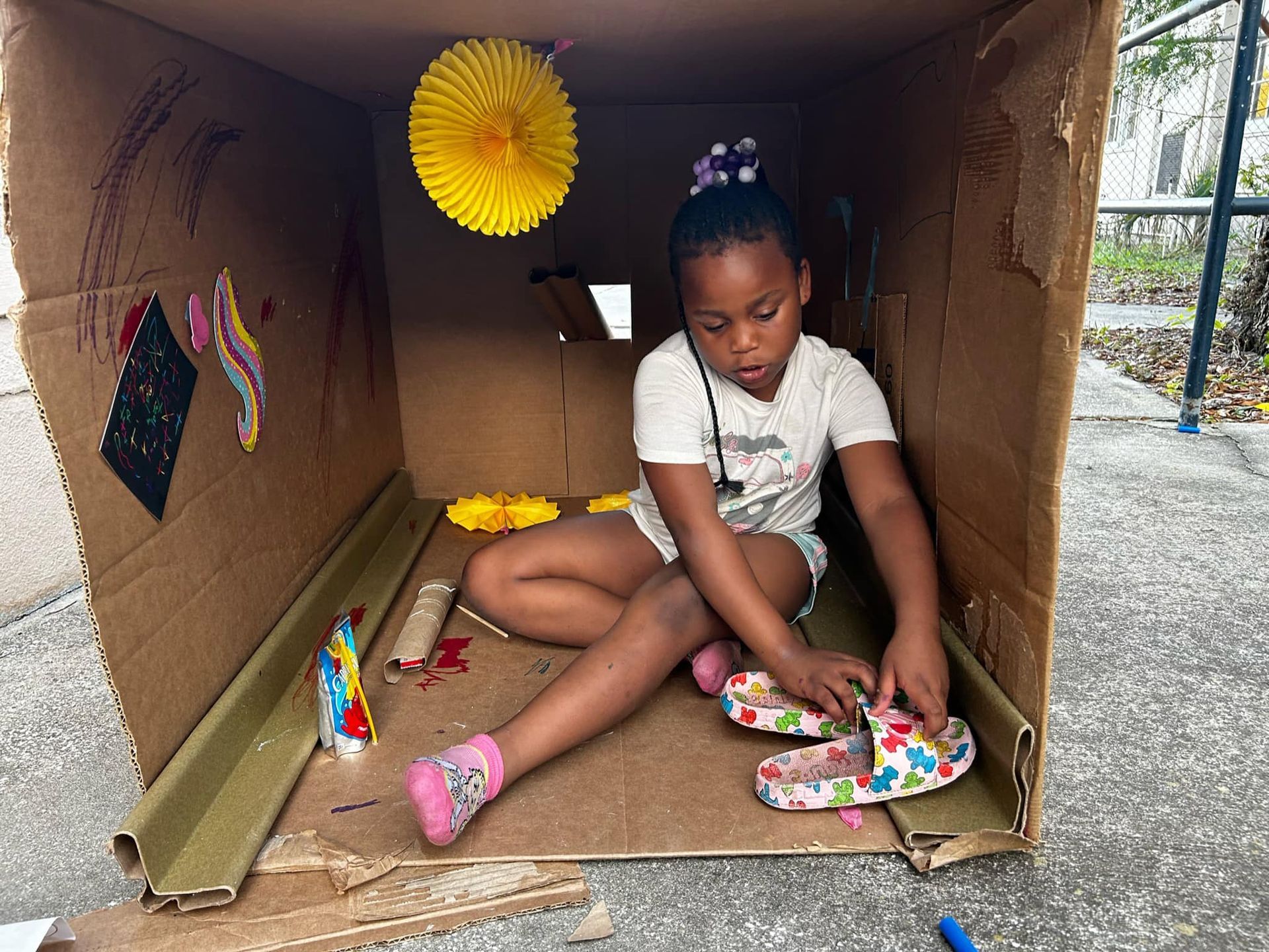 A little girl is sitting in a cardboard box playing with her shoes.