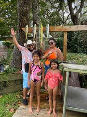 A family poses outdoors by a rack holding four fish, with the adults waving and smiling.