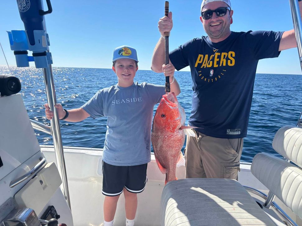 A child and an adult smile while holding a large red snapper fish on a boat on the open ocean.