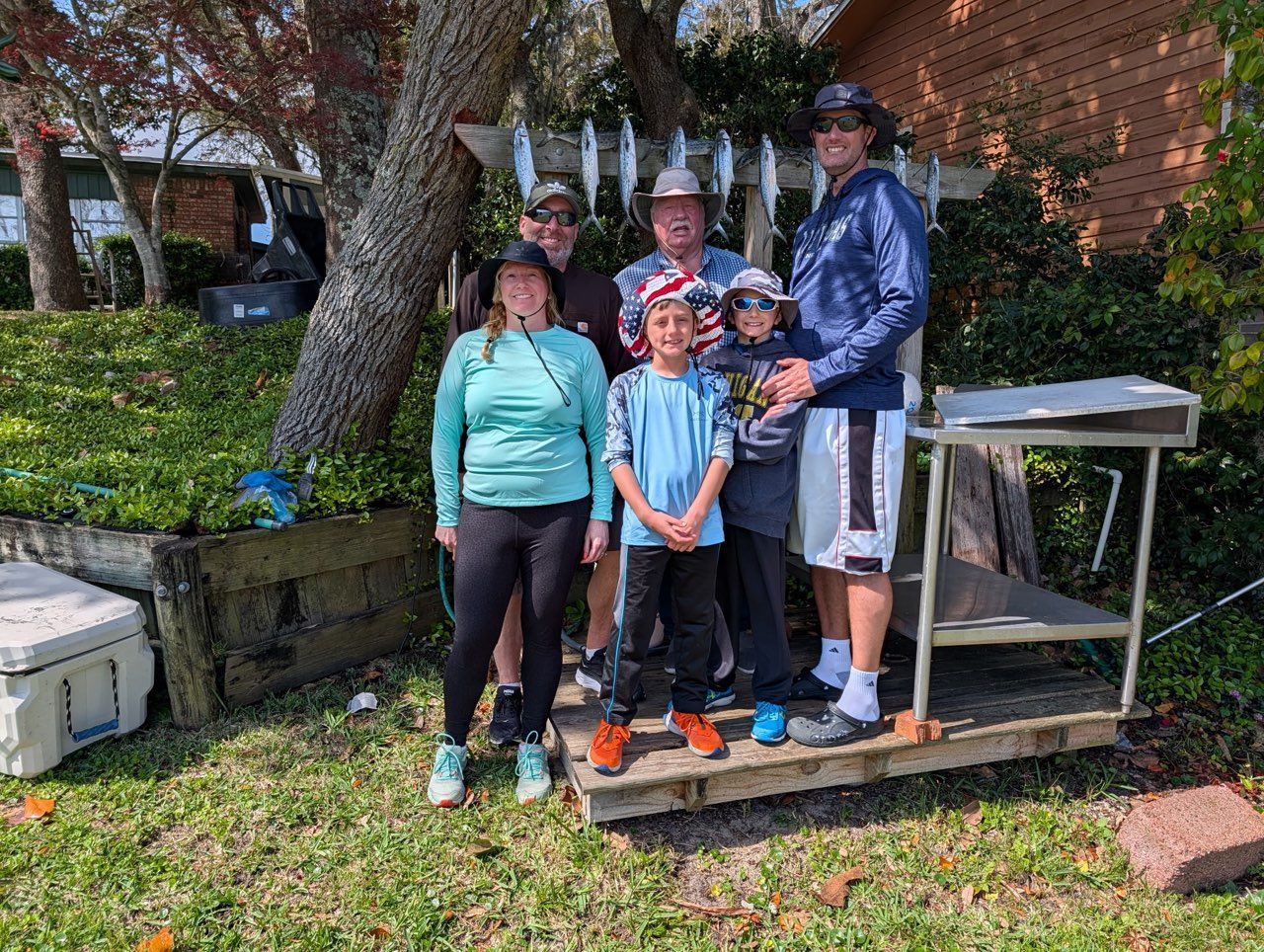 A family posing outdoors on a wooden platform near a rack of caught fish, with a cooler and trees in the background.