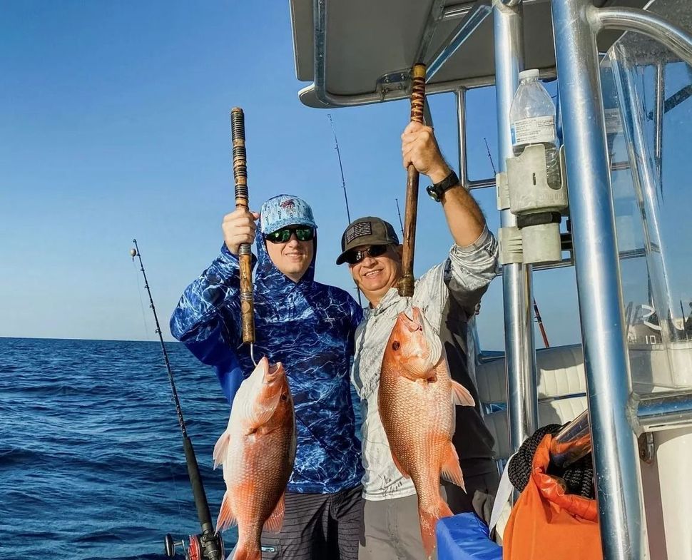 Two people on a boat hold up large red snapper fish caught while deep-sea fishing on a sunny day.