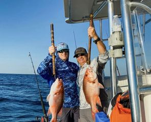 Two people on a boat holding up two large red snappers caught while deep-sea fishing under a clear blue sky.