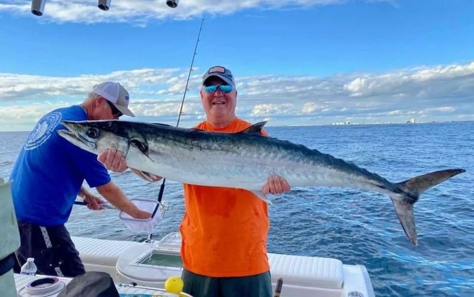 Two people on a boat holding a large king mackerel fish above the water on a sunny day.