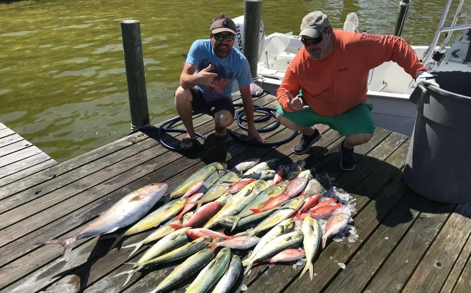 Two people crouching on a dock next to a large pile of freshly caught fish, smiling and gesturing at the camera.