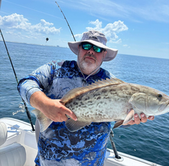 A person wearing a sun hat and sunglasses holds a large, spotted fish on a boat on the ocean under a blue sky.