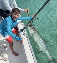 A smiling person on a boat holds a fishing rod, bringing a large silver fish to the surface of the clear, turquoise water.