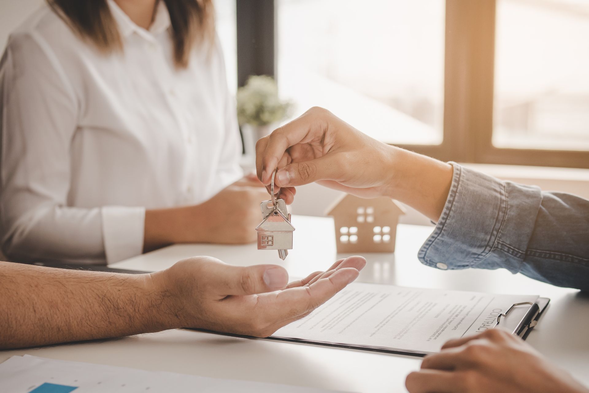 Hands exchanging house keys over a document, with a small wooden house model on a desk in the background.