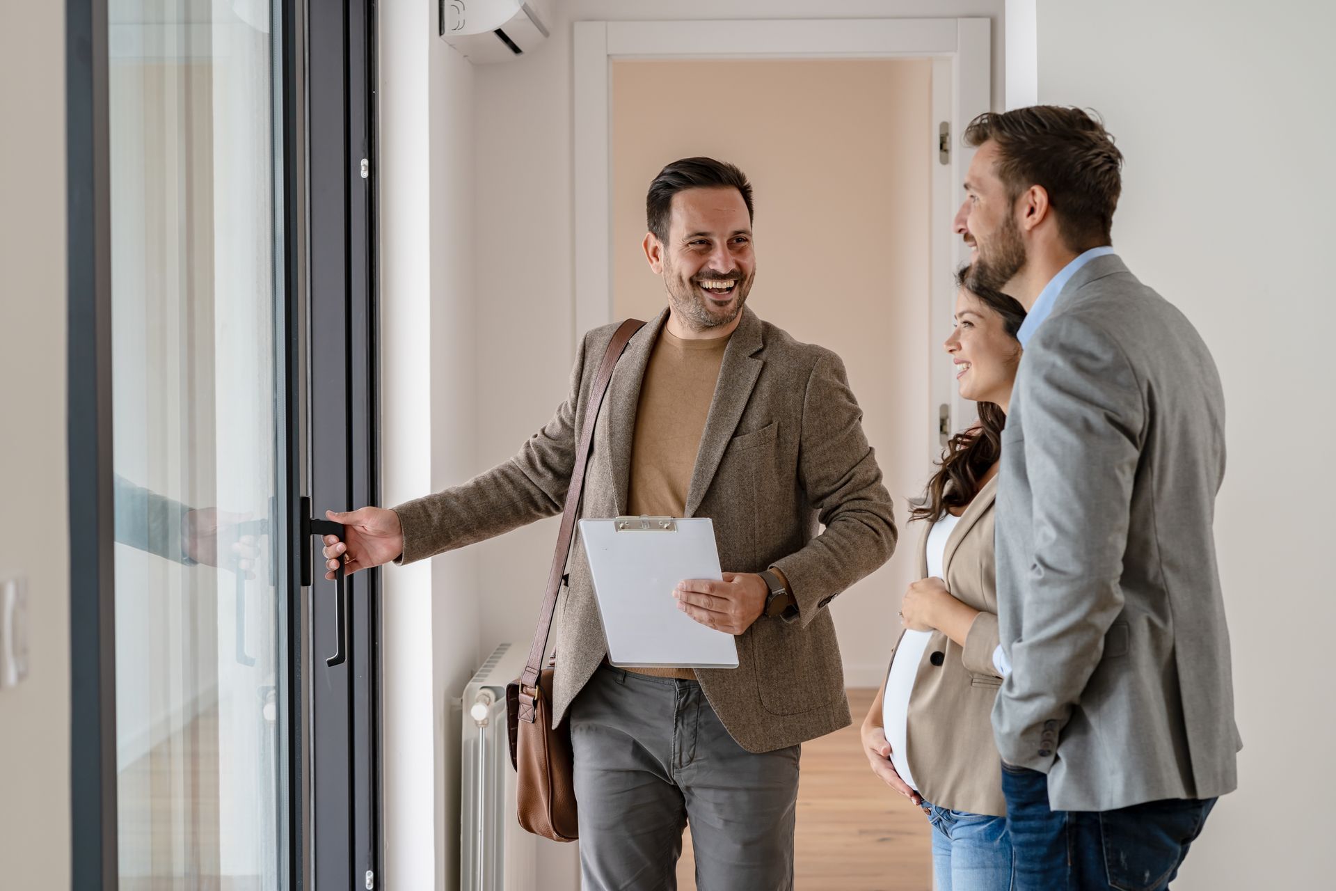 A real estate agent gestures to a glass door while showing a home to a couple in a bright, modern interior.