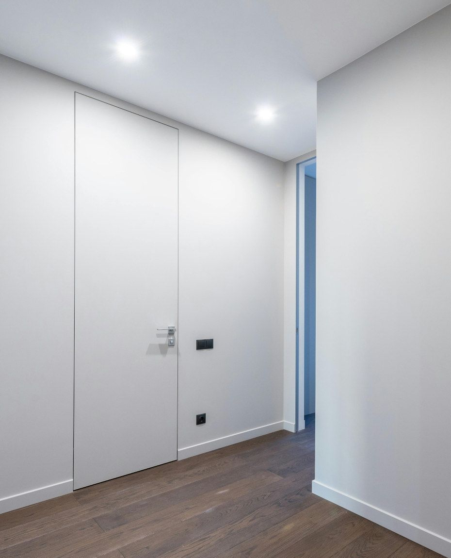 White hallway with dark wooden floor, a closed door, and recessed lighting. Another open doorway is visible to the right.