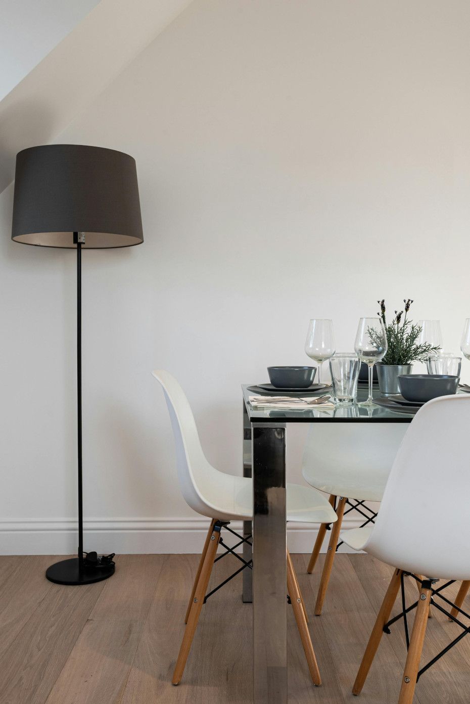 Dining area with white chairs, a glass-top table, and a tall black lamp against a white wall.