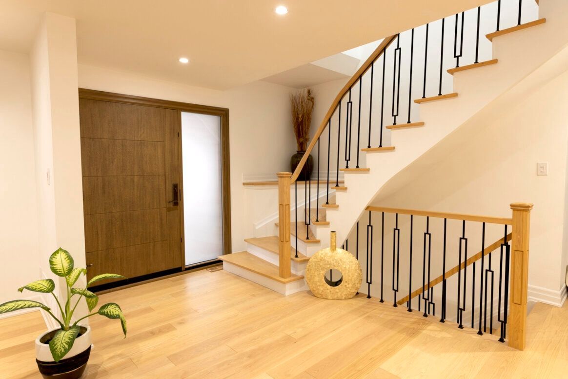 Light-filled entryway with wood door, staircase, and decorative vase. Blonde wood floors and white walls create a modern interior.