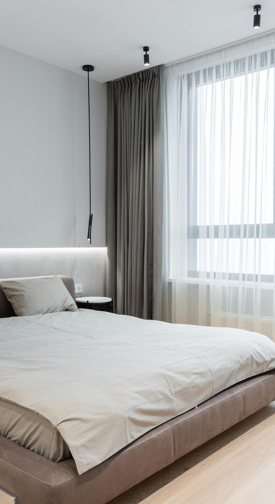 Modern bedroom with a bed, window, and gray curtains. The bed has gray bedding and a leather headboard.